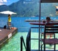 Outdoor area by a lake with wooden deck, lounge chairs, umbrella, and a diving board with a person jumping into the water. Scenic mountain view in the background.