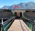 A wooden boardwalk leads to a building labeled "Lido Riva Caccia" with flags and a scenic mountain view in the background.