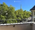 Balcony with metal railing overlooking a street lined with trees and buildings, including a church spire in the distance.