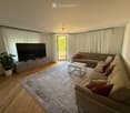 Modern living room with a large sectional sofa, coffee table, flat-screen TV, and a potted plant. The room features wooden flooring, large windows with white curtains, and access to a balcony.