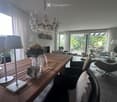 Modern living room with a wooden dining table, sofa, chairs, chandelier, lamps, and a view of the garden through large windows.