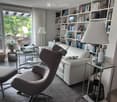 Modern living room with a white leather sofa, armchair, glass coffee table, bookshelf filled with books, and a large window with a view of the garden.