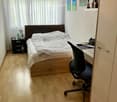 A small, well-lit bedroom with a wooden floor, a bed with white bedding, a nightstand, a desk with a chair, and several books and decorative items on shelves.