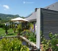Well-maintained garden with a pond, various plants, flowers, and a modern house in the background. Features include a patio umbrella, decorative statues, and a scenic mountain view.