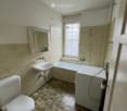 Bathroom with beige tiled walls, a bathtub, a toilet, a sink, a mirror, and a window with frosted glass. The floor is tiled with a dark, speckled pattern.