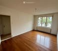 Empty room with wooden flooring, white walls, a window with a radiator below, and a door leading to another space.
