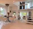 Modern, well-lit living and dining area with a round dining table, white chairs, a chandelier, and a staircase leading to a lofted bedroom area with a sofa and decorative items.
