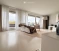 Modern living room with large windows and balcony access, featuring a white sofa, armchair, coffee table, floor lamp, and decorative vase.