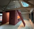 Loft-style room with exposed wooden beams, a staircase leading to an upper level, a compact kitchen area, and large windows allowing natural light.
