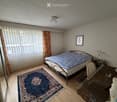 A furnished bedroom with a bed, a chair, a table, a picture frame, and a lamp. The room features wooden flooring, a large window with curtains, and a blue rug.