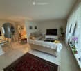 Bright living room with tiled flooring, large windows with white curtains, and a mix of seating including a sofa, armchair, and dining table with chairs. A television is mounted on a white stand, and decorative elements such as lamps, a mirror, and artwor