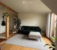 Modern living room with wooden flooring, green sectional sofa, ottoman, side table, hanging guitar, and plants.