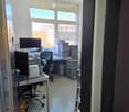 Office space with a desk, chair, screen, keyboard, and several stacked crates. The room has large windows and a door leading to another area.