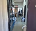 A clean, well-lit hallway with metal shelving units on the left side and various equipment and supplies on the right side, including a cart, a computer setup, and a white machine with papers attached.