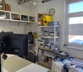 Workshop or office space with a table, keyboard, chair, shelves, clock, screen, printer, and tools.
