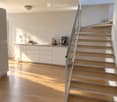 Modern, minimalist kitchen area with white cabinets, wooden flooring, and a staircase with wooden steps and a glass railing. A mixer, bowl, and bottles are placed on the counter.