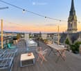 Rooftop terrace with wooden decking, string lights, a small table with a bowl, chairs, a bench, and a barbecue, overlooking a cityscape with a prominent church tower.