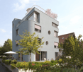Modern, multi-story building with a sleek, contemporary design, featuring large windows, a rooftop terrace, and surrounding greenery.