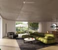 Modern living room with a green sectional sofa, black leather armchair, white coffee table, dining area with wooden chairs, pendant lights, and large windows overlooking a garden with an umbrella.