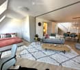 Modern, open-concept living and bedroom area with a grey sofa, wooden coffee table, bed with red bedding, small round table with a plant, and a dining area with chairs and a table.