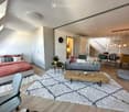 Modern, open-concept living and sleeping area with a grey sofa, wooden coffee table, dining table with chairs, bed with red bedding, and a potted plant on a side table.