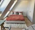 A cozy attic bedroom with a slanted ceiling and skylights, featuring a bed with a red blanket and decorative pillows, a wooden chair, a small table, and two fluffy rugs on the floor.