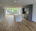 Open-plan kitchen and living area with light wood flooring, white island, grey cabinets, and large windows offering a view of greenery outside.