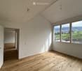 Empty room with wooden flooring, white walls, large windows offering a scenic view of green fields and mountains.