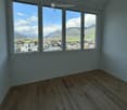 Empty room with wooden flooring and large windows offering a scenic mountain view of a town and surrounding landscape.