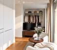 Modern living room with a white sofa, wooden coffee table, flat-screen TV, and a wardrobe with hanging clothes.