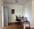 A furnished room with a wooden desk, office chair, lamp, and various items on the desk including a laptop, books, and a speaker. There is a handbag on the floor, a mirror on the wall, and a patterned rug on the floor.