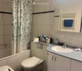 Modern bathroom with a toilet, sink, mirror, and cabinet. The sink has a faucet and is topped with a countertop holding various toiletries. A shower curtain with a leafy pattern is visible, and a towel rack with a blue towel is mounted on the wall.