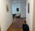 A narrow hallway with wooden flooring, white walls, and a large patterned rug. There are framed pictures on the walls, a small seating area visible at the end, and some bags placed on the floor.