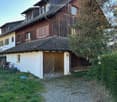 Two-story house with wooden exterior, tiled roof, and garage. The house features multiple windows with shutters and is surrounded by greenery.