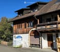 Two-story house with a tiled roof, wooden shutters, and a stone archway entrance. The building features a balcony on the upper floor and a signboard in front.