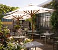 tables and chairs set up under umbrellas on a wooden deck with a view of a garden and car in the distance