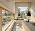 Modern kitchen and dining area with large windows overlooking a garden, featuring a dining table, chairs, pendant lights, and a countertop with a bowl and vase.