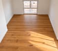 Empty room with light wooden flooring and white walls, featuring large windows allowing natural light to enter.