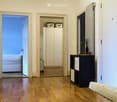 A modern hallway with wooden flooring, a cream-colored sofa, a black cabinet with drawers, and picture frames on the wall. An open doorway leads to a bedroom with a bed visible in the background.