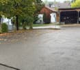 Empty paved area with scattered leaves, surrounded by trees and houses.