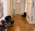 A modern hallway with herringbone wooden flooring, white walls, and two dark green chairs flanking a small round table with a vase of flowers. A closed door leads to another room.