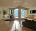 Bright living room with wooden flooring, large windows offering a sea view, a grey sofa, a fireplace, a TV, and a small coffee table.