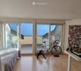 Bright room with wooden flooring, large sliding glass doors leading to a balcony, a bed, a desk with a laptop and lamp, a bicycle, and an exercise bike.