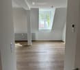 Empty room with wooden flooring, white walls, and a window with a view of greenery. The room features a radiator and a door with a handle.