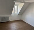 Empty attic room with wooden flooring, white walls, and a single window with a slanted roof design. A radiator is visible against one wall.