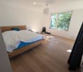 Bright bedroom with wooden flooring, large window, bed with white quilt and blue pillow, nightstand, fan, and backpack near the window.