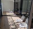Modern balcony with tiled floor, large glass sliding doors, two white cushioned chairs, and a small shelf visible through the glass.
