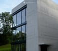 Modern, minimalist concrete building with large glass windows and a raised ground floor entrance.