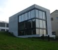 Modern concrete building with large glass windows, situated on a raised ground floor with a grassy slope leading up to it. Adjacent buildings and parked cars are visible in the background.