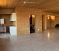 Open-plan space with exposed wooden ceiling and walls, featuring a small kitchen area with a refrigerator, sink, and cabinets, and a large open area with wooden walls and a polished concrete floor.